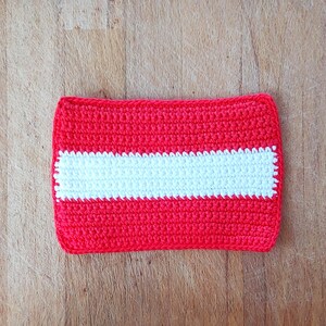 May include: A rectangular, crocheted item featuring a vibrant red background with a horizontal white stripe in the center. The handmade piece showcases a textured stitch pattern, likely a small pouch or decorative item.