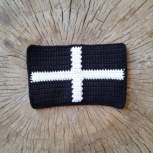 May include: A black and white crocheted square with a white cross design. The square is made of yarn and has a textured surface.