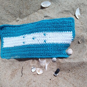 May include: A handmade crochet item in shades of turquoise and white, resting on sand with seashells. The rectangular piece features a turquoise border and a central white stripe with star-shaped cutouts. The texture is visible, showcasing the yarn's weave.