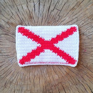 May include: A crocheted square with a red cross design on a white background. The cross is made of red yarn and is centered on the square.