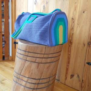 May include: A large, handmade, crochet duffel bag in a periwinkle blue. The bag features a rainbow design on one end and green and yellow straps. The bag is sitting on a wooden stump.