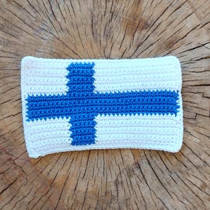 May include: Crocheted Finnish flag in white and blue, with a blue cross on a white background. The flag is lying on a wooden surface.