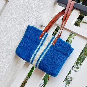 Könnte beinhalten: Blaue Tragetasche mit zwei weißen Streifen in der Mitte. Sie hat braune Ledergriffe und hängt an einem weißen Zaun. Die Tasche ist aus einem weichen, strukturierten Material gefertigt.