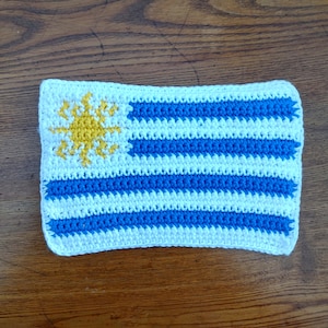 Pode incluir: Uma bandeira uruguaia feita de crochê. A bandeira é azul e branca com um sol amarelo no canto superior esquerdo.