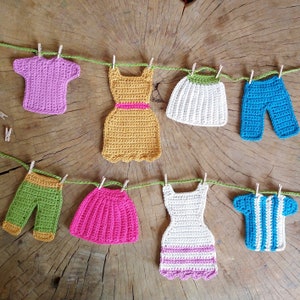 May include: A string of crocheted clothing items hanging on a clothesline. The items include a pink shirt, a yellow dress, a white skirt, a blue pair of trousers, a green pair of trousers, a pink skirt, a white dress with pink trim, and a blue and white striped shirt.