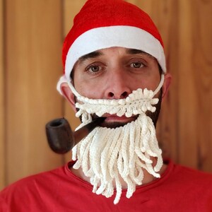 May include: A red Santa hat with a white brim and a crocheted white beard. A brown pipe is held in the mouth. The person is wearing a red shirt. The background is a wooden wall.