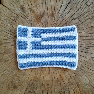 Pode incluir: Bandeira grega de crochê em azul e branco, com uma cruz branca sobre fundo azul. A bandeira está sobre uma superfície de madeira.