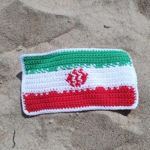 Könnte beinhalten: Gehäkelte iranische Flagge mit horizontalen Streifen in Grün, Weiß und Rot. Der weiße Streifen hat ein rotes Emblem in der Mitte. Die Flagge ist rechteckig und liegt auf Sand.