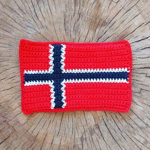 May include: A crocheted Norwegian flag made with red, white and blue yarn. The flag is square with a white cross on a red background. The cross has a blue border.