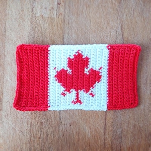 May include: A crocheted Canada flag with a red maple leaf on a white central panel, framed by red borders. The handmade flag is placed on a wooden surface. The flag represents Canadian national identity.