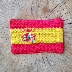 May include: A crocheted Spanish flag with red, yellow, and gold colors. The flag features a coat of arms in the center.