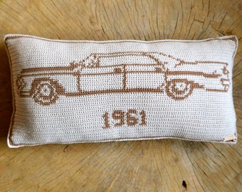 May include: A beige crocheted pillow with a brown outline of a 1961 car. The pillow has the year "1961" stitched in brown on the front.