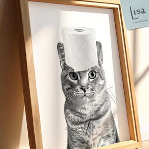 Op de afbeelding: Een zwart-wit afdruk in een lijst van een tijgerkat met een rol toiletpapier op zijn hoofd. Het kunstwerk zit in een houten lijst en heeft een label met "lisa" en "Lisa's Artwork". De afdruk leunt tegen een muur.