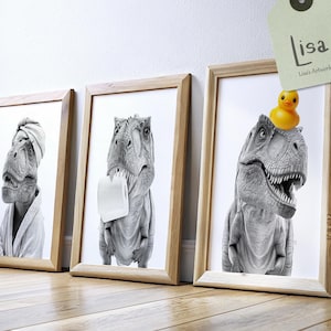 May include: Three framed black and white dinosaur art prints. One dinosaur wears a towel on its head and a robe. Another holds toilet paper. The third has a rubber duck on its head. The frames are light brown.
