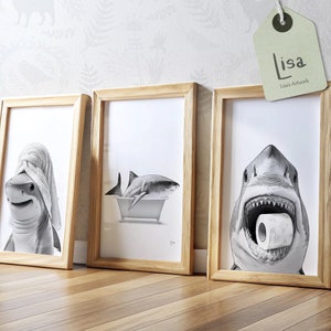 May include: Three framed black and white illustrations of sharks. The first shows a shark wearing a towel on its head. The second shows a shark in a bathtub. The third shows a shark with its mouth open holding a roll of toilet paper. The frames are made of light wood.