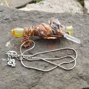 May include: A copper wire wrapped pendant featuring a clear quartz crystal, pink and white stones, and a yellow glass tube. The pendant is attached to a silver snake chain necklace.