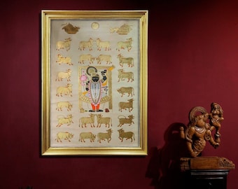 Puede incluir: Una pintura enmarcada de una deidad hindú con un marco dorado y un fondo blanco. La deidad está rodeada de 24 vacas, todas en oro. La pintura está colgada en una pared marrón.