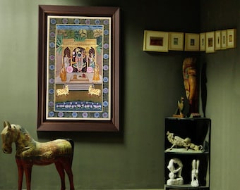 Puede incluir: Una pintura en miniatura india enmarcada con un marco de madera marrón oscuro. La pintura representa una escena religiosa con figuras y animales en colores vibrantes. Un caballo de madera sobre ruedas está en primer plano. Otros objetos decorativos están en el fondo.