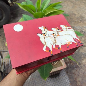 May include: A red wooden box with a painted design of white cows. The box has a white moon on the front and a gold trim around the edges.