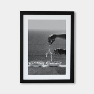 May include: Black and white framed photograph of two hands twirling pasta with a fork, above two bowls of pasta. The background shows a body of water and a hazy sky. The image is a close-up shot.