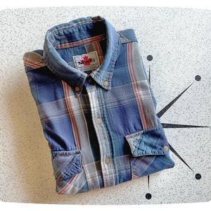 Puede incluir: Una camisa de franela a cuadros azules y rojos con cuello abotonado y un bolsillo de parche en el pecho izquierdo. La camisa tiene una etiqueta que dice "NAVS" con un logotipo de hoja de arce.