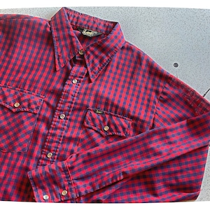 Puede incluir: Una camisa de manga larga a cuadros rojos y azules con cuello de punta y dos bolsillos en el pecho. La camisa tiene el logotipo de la marca Lee en el bolsillo y el cuello.