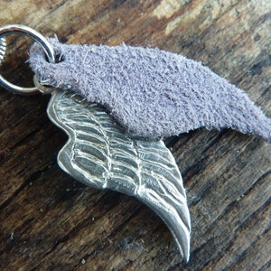 May include: A silver pendant featuring a detailed wing design, attached to a silver chain. The pendant is topped with a small piece of purple suede.