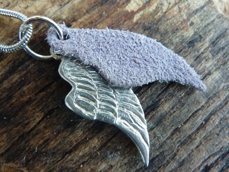 May include: A silver pendant featuring a detailed wing design, attached to a silver chain. The pendant is topped with a small piece of purple suede.