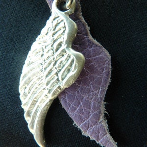 May include: A silver wing pendant with a textured surface, attached to a purple leather cord.