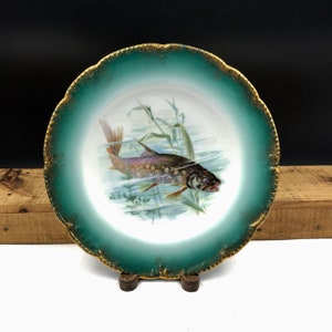 May include: A vintage porcelain plate with a green and gold rim. The center of the plate features a hand-painted image of a trout swimming in a stream.