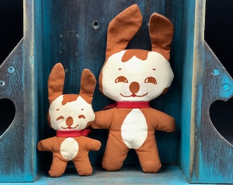 May include: Two brown and white fabric bunny dolls with red ribbon accents. The larger bunny is standing in front of the smaller bunny. Both bunnies have smiling faces.