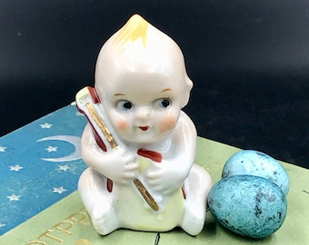 May include: A porcelain figurine of a baby holding a red and gold bow and arrow. The figurine is sitting on a book with a blue and white star pattern.