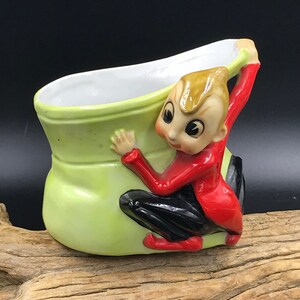 May include: A green ceramic boot planter with a small elf figurine holding onto the side. The elf is wearing a red jacket, black pants, and red shoes.