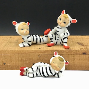 May include: Three ceramic figurines of children dressed in black and white zebra print pajamas. The figurines are in various poses, including lying down, sitting, and standing. The figurines are made of white ceramic with black stripes and have painted facial features.