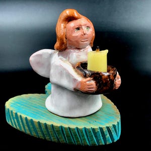 May include: A ceramic angel figurine with reddish-brown hair and a white robe, holding a dark brown bowl containing a small yellow candle. The angel is mounted on a heart-shaped base painted in a textured, vibrant turquoise blue color.