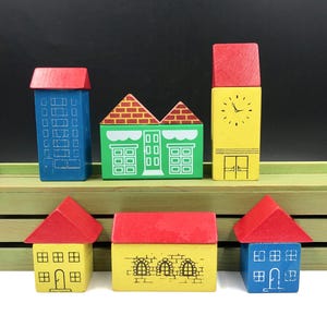 May include: A collection of colorful wooden toy blocks shaped like buildings. The set includes a blue skyscraper, a green shop, a yellow clock tower, and three houses with red roofs. The blocks are painted with details like windows and doors.