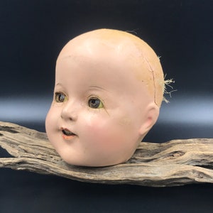 May include: A vintage porcelain doll head with painted blue eyes, a pink blush, and a slightly open mouth. The doll head is cracked and has a few strands of hair attached to the top.