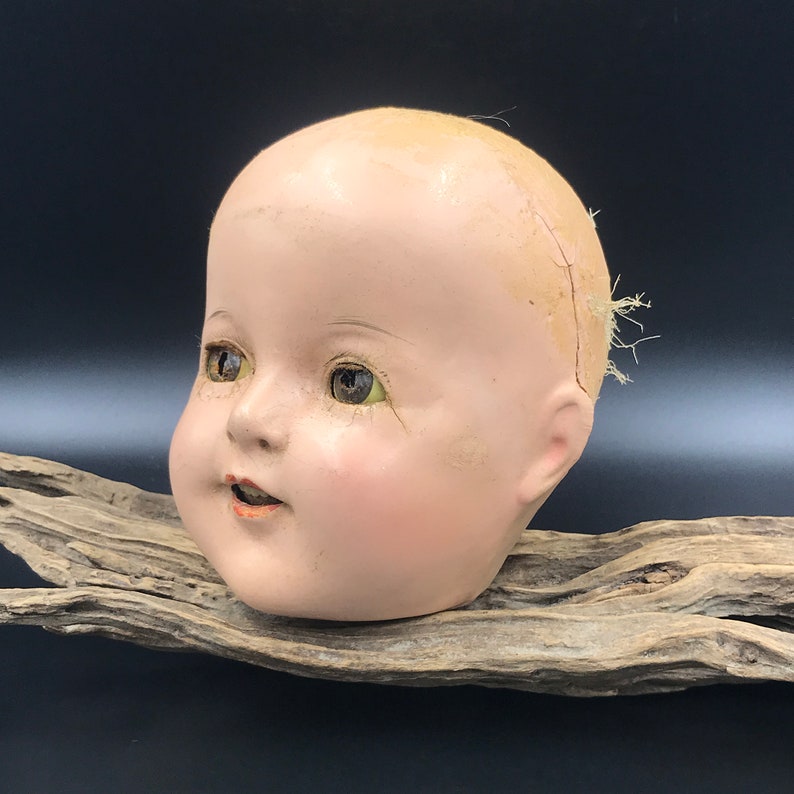 May include: A vintage porcelain doll head with painted blue eyes, a pink blush, and a slightly open mouth. The doll head is cracked and has a few strands of hair attached to the top.