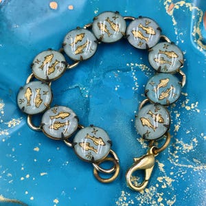 May include: A vintage bracelet featuring light blue circular links with gold fish designs. The bracelet has a gold-tone metal frame and clasp. The fish are in a swimming pose. The bracelet is on a blue surface.