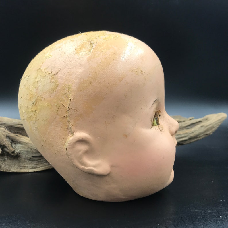 May include: A vintage doll head with cracked and peeling paint. The doll has green glass eyes and a painted smile. The head is made of a hard, plaster-like material.