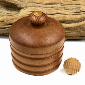 May include: A round, medium-brown wooden lidded container with horizontal ribbed detailing on the sides and a small, circular knob handle on top. Beside the container sits a separate, small, round wooden piece with a textured grid pattern.