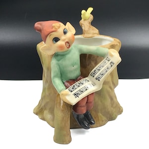 Puede incluir: Una figurita de cerámica de un elfo cantando desde un tocón de árbol con un pájaro amarillo posado en una rama arriba. El elfo lleva un gorro rojo, un suéter verde y pantalones marrones. El elfo sostiene una hoja de música con notas musicales.