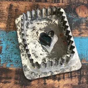 May include: A vintage metal cookie cutter set. The set includes a rectangular cutter with a scalloped edge and a small heart-shaped cutter. The metal has a worn, aged appearance, with signs of use and wear. The background is a rustic wooden surface.