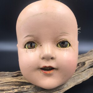 May include: A vintage porcelain doll head with painted green eyes, a closed mouth, and a rosy complexion. The doll head is missing its hair and has some wear and tear.