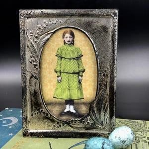 May include: A rectangular silver-toned metal picture frame with an oval center opening featuring embossed lily of the valley floral details. Inside the frame is a vintage-style portrait of a person wearing a lime green polka dot dress with a ruffled collar.