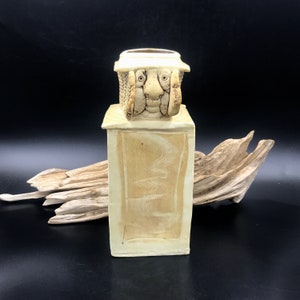 May include: A ceramic vase with a whimsical face and a textured, cream-colored body. The vase is decorated with a wavy pattern.