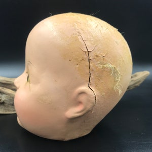 May include: A vintage doll head made of porcelain with a crack down the side. The head is painted in a light pink color and has a detailed face with painted eyes and a slightly open mouth. The doll's ear is visible on the side of the head.