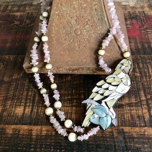 May include: A handcrafted necklace featuring a parrot pendant made of iridescent shell pieces. The necklace has strands of small, light purple stones and cream-colored beads with gold-colored accents. The parrot pendant is perched on a branch with blue leaves.