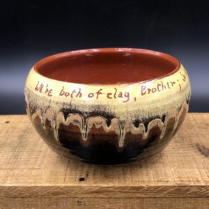 May include: A brown ceramic bowl with a yellow glaze and black accents. The bowl has a handwritten inscription that reads "We're both of clay, Brother, we".