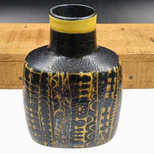 May include: A ceramic vase with a textured black finish and a yellow rim. The body features vertical bands of intricate, etched golden-yellow geometric patterns and floral motifs. The vase has a rounded, slightly flattened shape with a narrow neck.